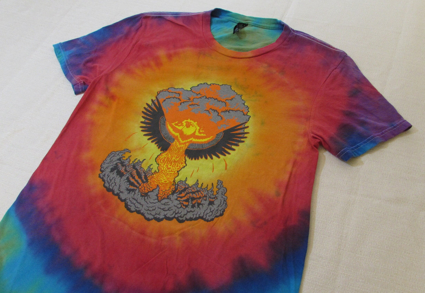 A phoenix rising out of a mushroom cloud on a tie dye shirt with a white background