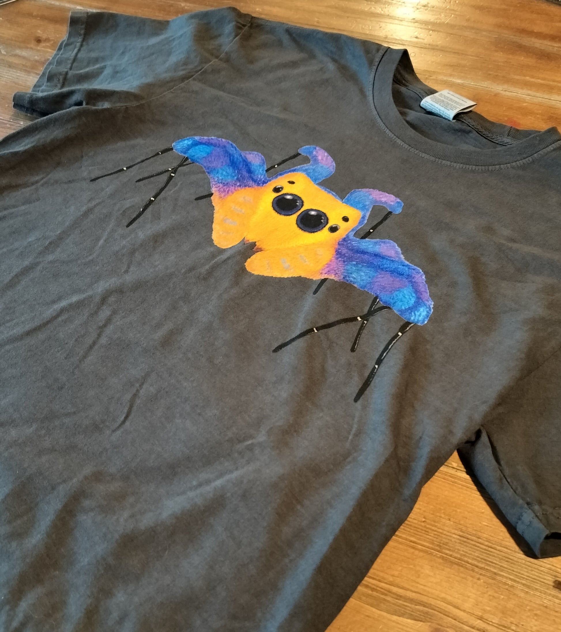 Gray t-shirt with a colorful jumping spider design on a wooden surface