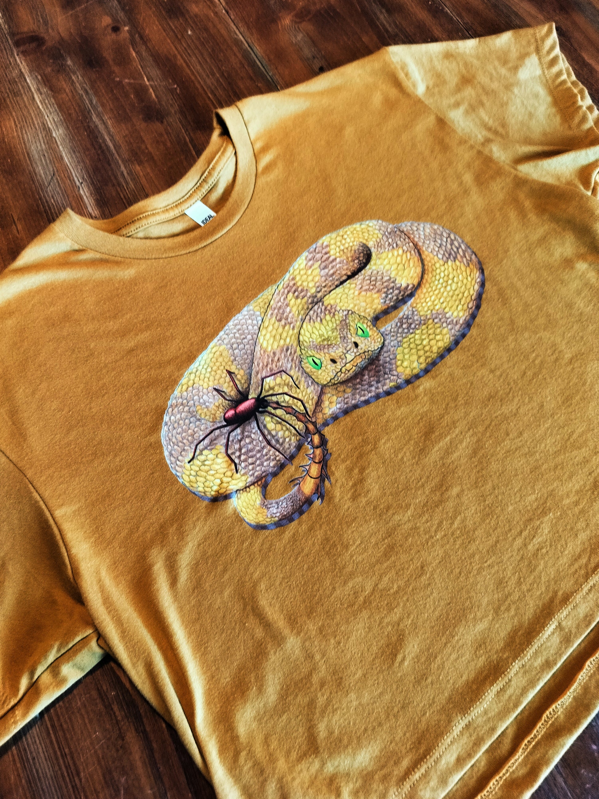 Gold t-shirt with a snake and spider graphic on a wooden surface