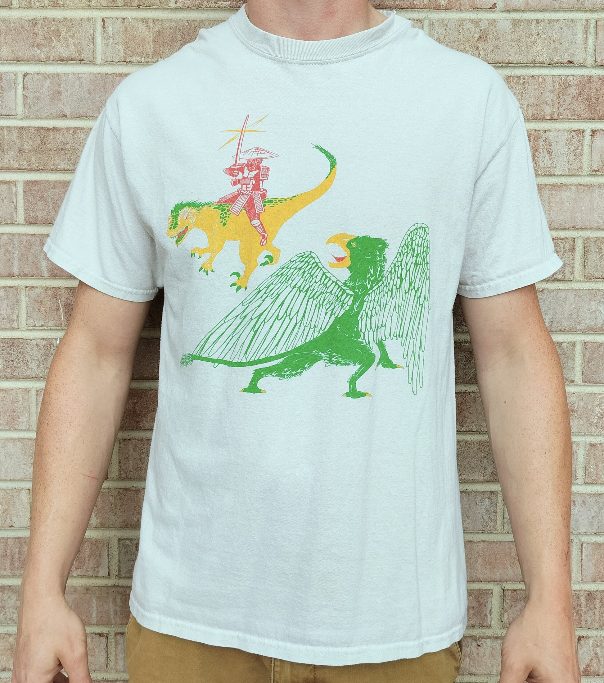 A men's t-shirt with an illustration of a samurai, raptor, and griffon in front of a brick background 