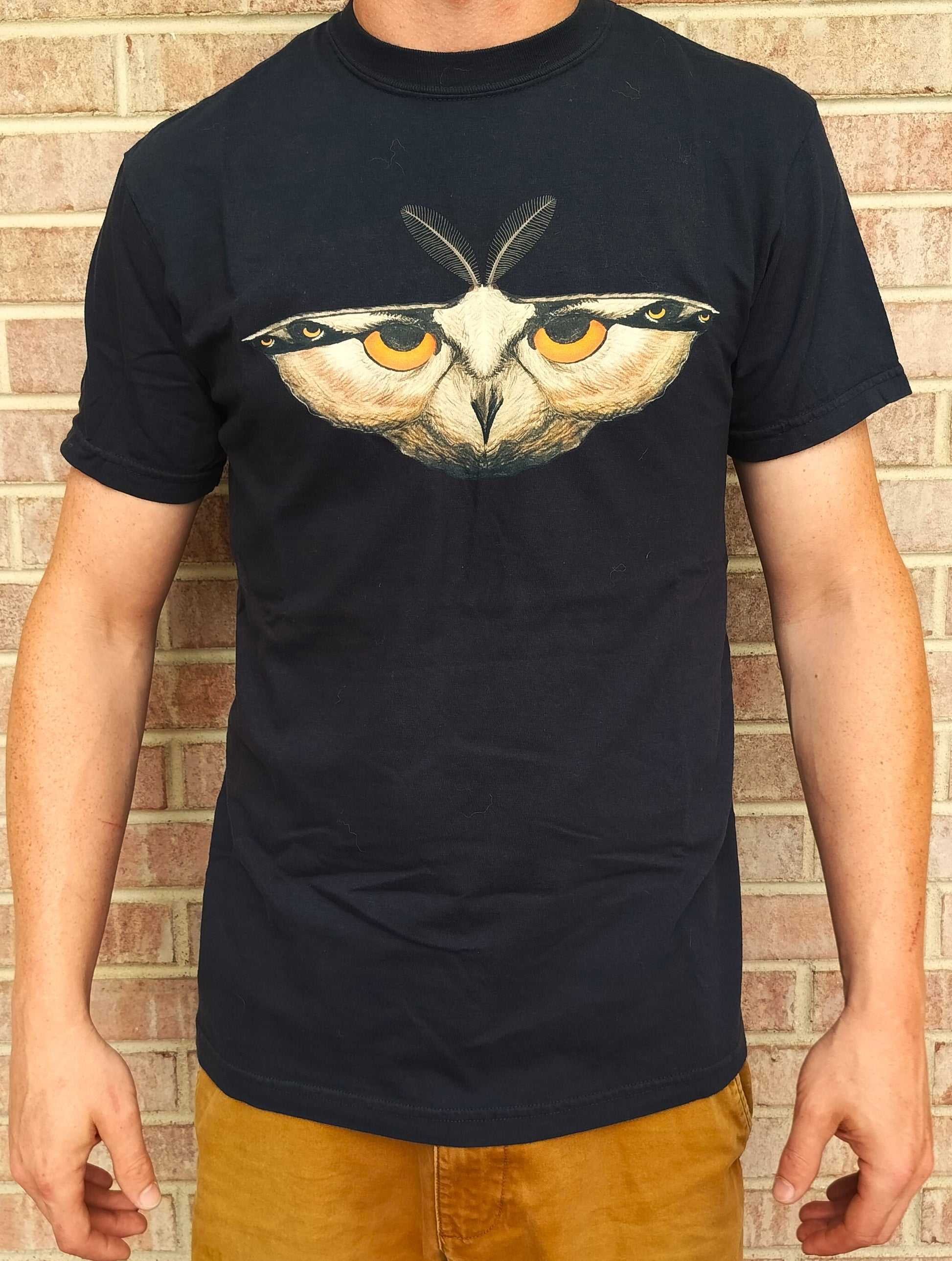 A person wearing a navy-colored T-shirt with a front print of a stylized moth, specifically an Antheraea polyphemus moth, with prominent eye spots.